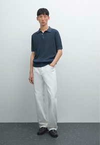 Polo shirt navy a maniche corte con un motivo testurizzato, abbinato a pantaloni bianchi a gamba dritta e mocassini neri, mani in tasca.