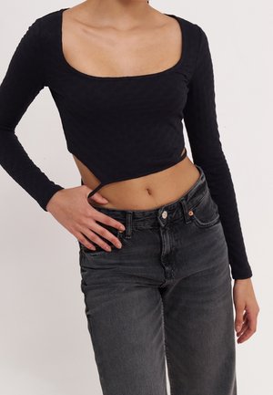Black long-sleeve top with a square neckline, textured fabric, cropped cut, and side tie detail, paired with high-waisted black jeans.