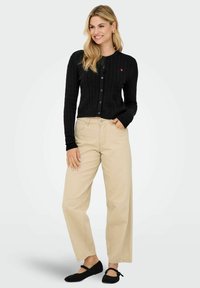 Cardigan nero a maglia grossa con bottoni e un piccolo logo a forma di cuore rosso. Abbinato a pantaloni larghi beige chiaro e ballerine nere.