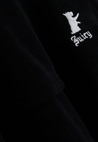 Μαύρο ύφασμα με λευκό κεντητό σχέδιο που θυμίζει σκύλο πομά εκ πάνω από τη λέξη "Juicy" σε στυλιζαρισμένη γραφή.