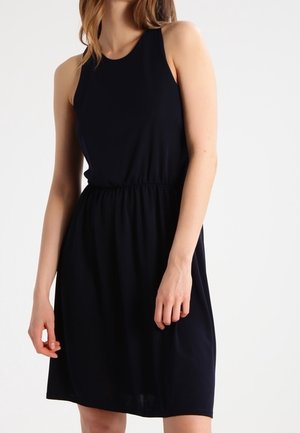 Vrouw die een mouwloze, knielange zwarte jurk met een geplooide taille draagt, staand tegen een effen lichte achtergrond.