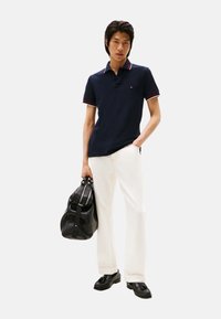 Polo azul marino con cuello a rayas rojas y blancas; combinado con pantalones blancos y bolso duffle de cuero negro. Zapatos negros completan el conjunto.