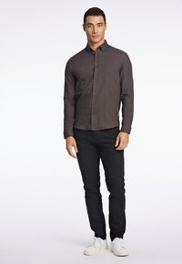 Camicia marrone con bottoni, maniche lunghe e bottoni scuri, abbinata a pantaloni neri e sneakers bianche, in piedi contro uno sfondo semplice.