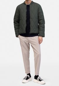 Pikowana zielona kurtka ze stójką, zestawiona z granatowym swetrem, jasnobeżowymi spodniami i czarnymi trampkami z białymi podeszwami.