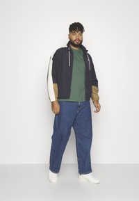 Marineblå og tan-fargeblokkerte jakker med hvite detaljer, glidelås-lukking og snorhette, kombinert med en grønn t-skjorte og løse blå jeans.