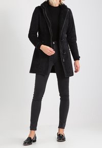 Vrouw die een zwarte schapenvachtjas draagt over een zwarte trui en zwarte skinny jeans met zwarte instappers, staande tegen een effen lichte achtergrond.