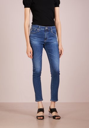 Kobieta w dopasowanych niebieskich dżinsach, czarnym topie z krótkim rękawem i czarnych sandałkach na otwarty palec z przezroczystymi słupkami, stojąca na beżowej podłodze.