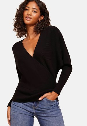 Pull noir côtelé enroulé avec un décolleté en V profond et des manches longues, associé à un jean en denim bleu, présentant une texture lisse et une coupe décontractée.