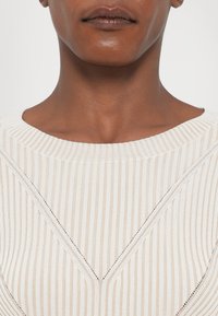 Tejpárot ribbelt pulóver, széles nyakkivágással és texturált átlós mintákkal, finom részletezéssel a széleken és enyhe fényláttal.