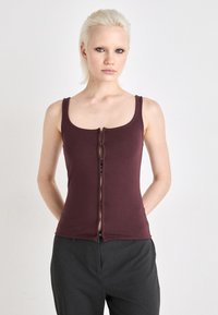 Top senza maniche in borgogna con scollatura a barchetta, chiusura frontale con zip e un design aderente realizzato in tessuto morbido e elasticizzato. Abbinato a pantaloni grigi.