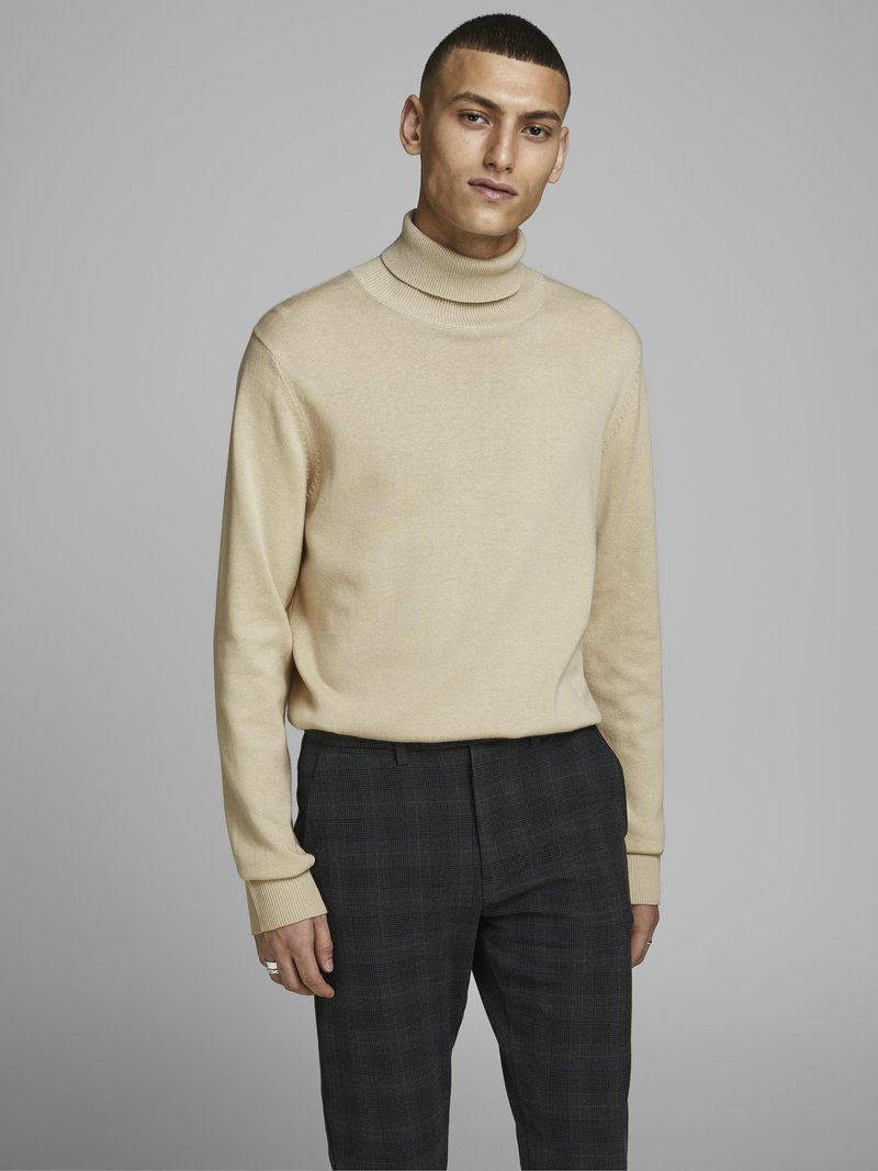 Pull à col roulé beige avec des poignets côtelés, associé à un pantalon sur mesure à carreaux sombres. Le tissu semble doux et léger.
