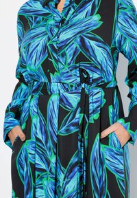 Svart klänning med livligt blått och grönt lövmönster, med krage, långa ärmar och ett knytbälte i midjan.