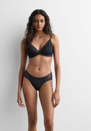 Kvinde med krøllet mørkt hår iført sort bh og matchende trusser med mesh-panel, stående mod en ensfarvet hvid baggrund.