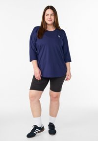 Marineblaues Sportoberteil mit dreiviertellangen Ärmeln, kombiniert mit schwarzen knielangen Shorts, weißen Knöchelsocken und schwarzen Schuhen mit weißen Streifen.