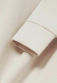 Svijetlosmeđi pamučni materijal s rubom na manžetama, s glatkom teksturom i ujednačenom bojom. Minimalistički dizajn bez uzoraka.
