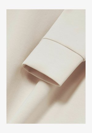 Gaiši beidzīgu kokvilnas audumu ar sarullētu piedurkni, kas izceļ gludu tekstūru un vienmērīgu krāsu. Minimālisma dizains bez rakstiem.