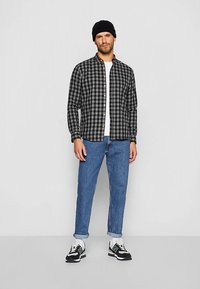 Black and gray plaid shirt, buttoned, white t-shirt underneath, blue jeans, black sneakers with white and green accents, wearing a black beanie.