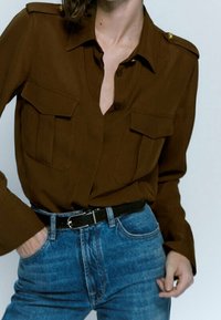 Brown button-up shirt with two chest pockets, featuring gold accents on the shoulders, paired with blue denim jeans and a black belt.