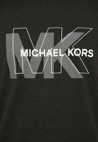 Čierna tkanina s prekrývajúcimi sa veľkými šedými a obrysovými bielymi písmenami "MK" a textom "MICHAEL KORS" vycentrovaným v bielej farbe.