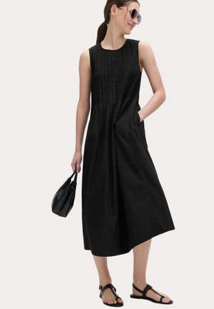 Vrouw draagt een mouwloze zwarte midi-jurk met plooien, zwarte sandalen, zonnebril, houdt een kleine zwarte handtas vast en staat voor een effen achtergrond.