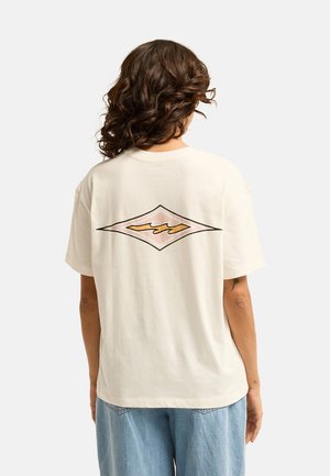 Kobieta o brązowych kręconych włosach do ramion nosi biały t-shirt z diamentowym, pomarańczowo-różowym wzorem fal na plecach oraz jasnoniebieskie dżinsy.