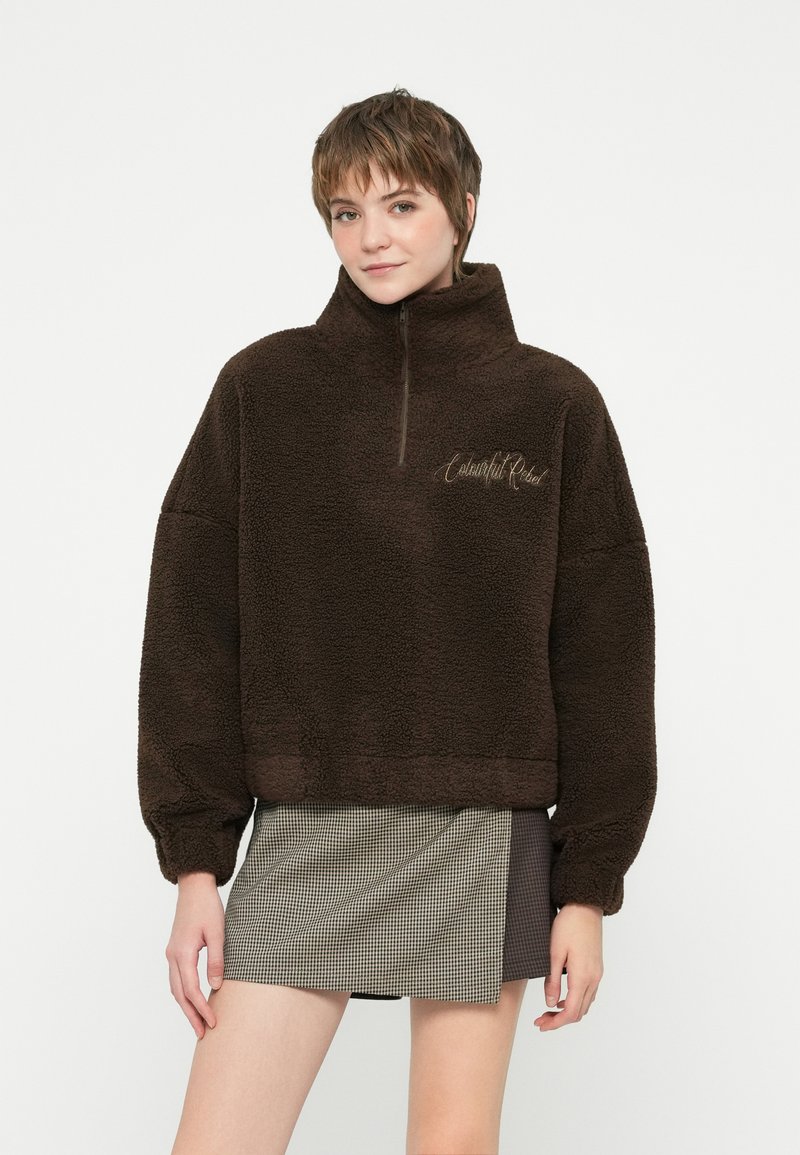 Pulover din fleece maro cu guler înalt, fermoar pe jumătate în față și manșete cu dantelă. Prezintă text brodat pe piept și țesătură texturată.