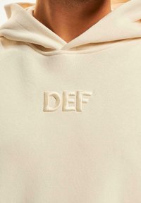 Hoodie beige en tissu texturé, avec un logo "DEF" brodé dans une teinte plus claire. La capuche est doublée d'un matériau doux.