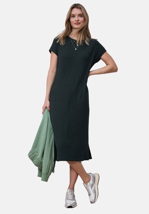 Kvinde stående iført en mørkegrøn midi-kjole og sølvfarvede sneakers, der holder en lysegrøn jakke i venstre hånd.