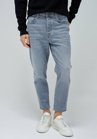 Jean en denim bleu clair avec une coupe décontractée, jambes fuselées et ourlets effilochés, associés à des baskets blanches. Le matériau semble doux avec un léger dégradé.