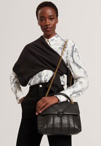 Zwarte leren handtas met een gestikt ontwerp en gouden kettingriem, vastgehouden tegen een patroonblouse en zwarte broek.