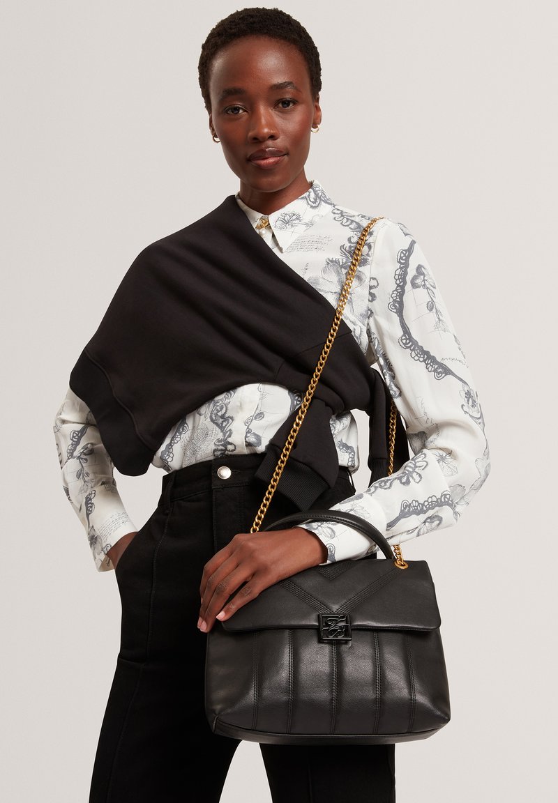 Zwarte leren handtas met een gestikt ontwerp en gouden kettingriem, vastgehouden tegen een patroonblouse en zwarte broek.