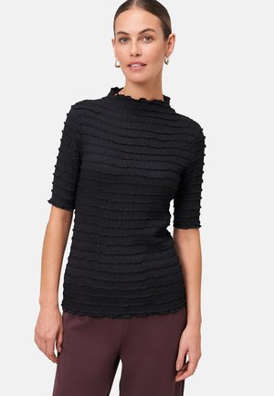 Femme portant un haut noir texturé à manches courtes avec des bordures ondulées et un pantalon bordeaux, se tenant devant un fond clair.