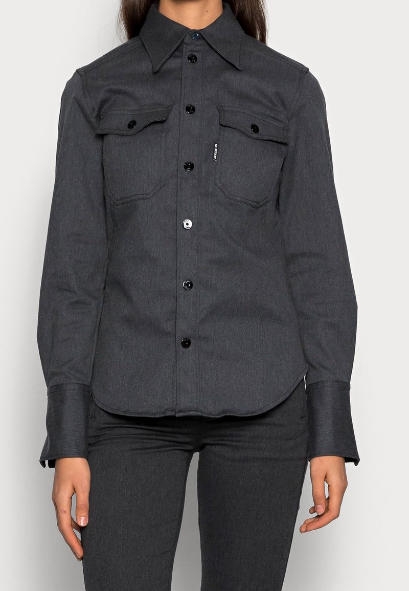 Femme portant une chemise boutonnée gris foncé avec deux poches poitrine et un pantalon gris foncé assorti, debout, les bras relâchés le long du corps.