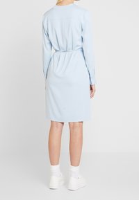 Vestido de manga larga azul claro con cintura ceñida, textura suave, cuello y puños abotonados. Llevado con zapatillas blancas y calcetines hasta el tobillo.
