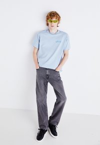 Hellblaues Baumwoll-T-Shirt mit "Levi's"-Logo, kombiniert mit schwarzen Jeans. Schwarze Sneakers und grüne Sonnenbrille runden den Look ab.
