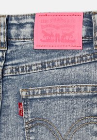 Le jean bleu Levi's présente une texture en denim délavé, un patch en cuir rose avec un texte embossé, et un onglet rouge sur la poche gauche.