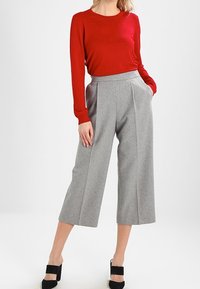 Haut rouge à manches longues et culottes grises taille haute. Les culottes présentent des plis et un design à jambes larges. Des escarpins à brides à la cheville noirs complètent la tenue.