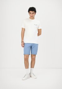 Camiseta blanca de algodón con un pequeño logotipo negro en el pecho, combinada con pantalones cortos azul claro arremangados y zapatillas blancas con cordones negros.