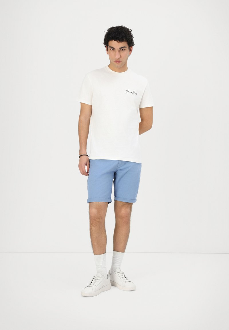 Weißes Baumwoll-T-Shirt mit einem kleinen schwarzen Logo auf der Brust, kombiniert mit hellblauen aufgerollten Shorts und weißen Sneakers mit schwarzen Schnürsenkeln.