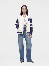 Γυναίκα με κρεμ και ναυτικό varsity μπουφάν πάνω από ριγέ πουκάμισο με τζιν παντελόνι καμπάνα και μυτερές καφέ παπούτσια, που στέκεται μπροστά σε απλό φόντο.
