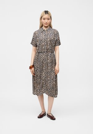 Femme aux longs cheveux blonds portant une robe midi à manches courtes imprimé léopard et des ballerines marron à lacets, debout sur un fond uni.