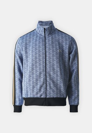 Blå zip-jacka i mjukt tyg, med geometriskt mönster, svarta muddar och nederkant, samt kontrasterande sidostreck. Lacoste-logotyp på bröstet.
