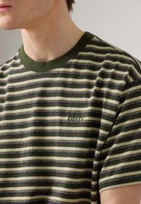 T-shirt de manga curta verde e creme às riscas, feita de algodão, com decote redondo e pequeno detalhe de logótipo no peito.