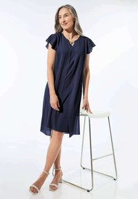 Navyblaues, knielanges Kleid mit kurzen, schwingenden Ärmeln, das eine leichte A-Linie und eine gefaltete Vorderseite aufweist. Paar beiger Blockabsatzsandalen.