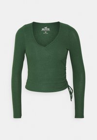 Grünes, geripptes Langarmoberteil mit V-Ausschnitt und Wickeldesign. Merkmale seitliche Raffung mit verstellbaren Bindebändern. Hergestellt aus einem strukturierten Strickmaterial.