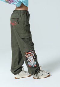 Olivgrön cargobyxor med elastisk midja, sidofickor och en broderad geisha-design på vänster ben. Matchas med vita sneakers.