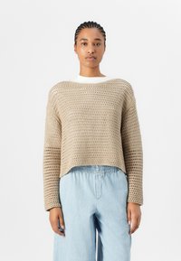 Beige Häkelpullover mit lockerer Passform, weitem Ausschnitt und langen Ärmeln. Kombiniert mit hellblauen, hoch taillierten, weiten Hosen.