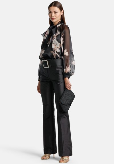 Blusa nera trasparente con motivi floreali, dettaglio a fiocco, abbinata a pantaloni neri in satin svasati e una clutch nera testurizzata. Tacchi con cinturini dorati.