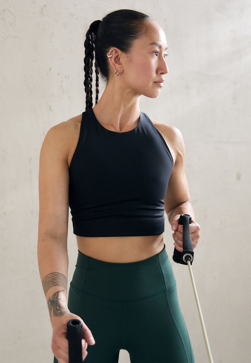 Čierny priliehavý športový crop top a vysoké zelené legíny. Osoba drží odporové gumy, čo ukazuje štýlový, atletický dizajn.
