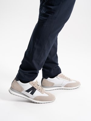 Person trägt schwarze Hosen und weiße Turnschuhe mit beigen Wildlederelementen sowie schwarze Knöchelsocken, steht vor einem weißen Hintergrund.